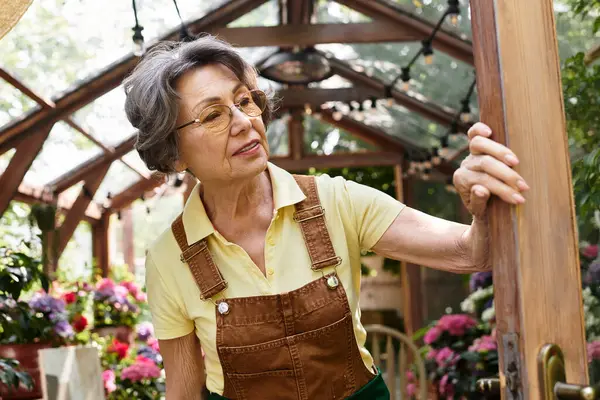 Beautiful senior woman smiles while opening the greenhouse door, surrounded by blooming plants. — Stock Photo