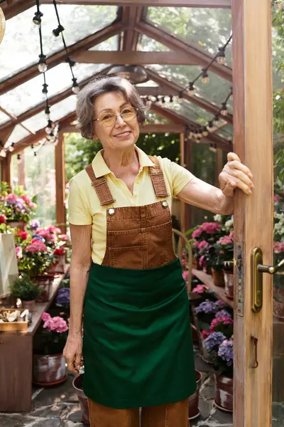 Smiling older woman opens the door of a greenhouse filled with colorful flowers in bloom. — стоковое фото