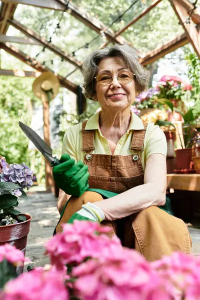 Joyful senior woman sits in a lush garden, skillfully nurturing blooming flowers with care. — Stock Photo