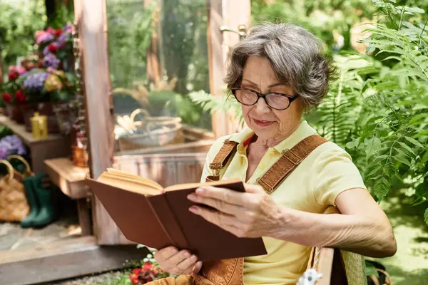 A senior woman with beautiful silver hair relaxes in her garden, lost in a book. — стокове фото