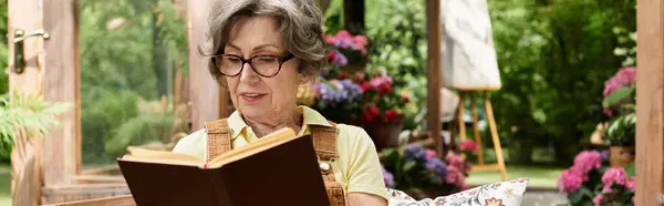 Beautiful senior woman sits relaxed in a colorful garden, absorbed in reading her book. — Stock Photo