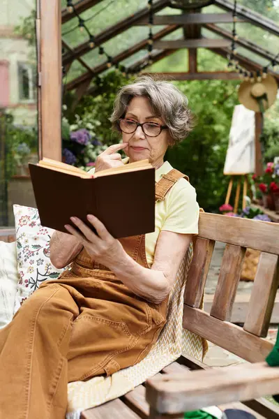 A senior woman sits peacefully on a bench in a lush garden, engrossed in a book. — Stock Photo