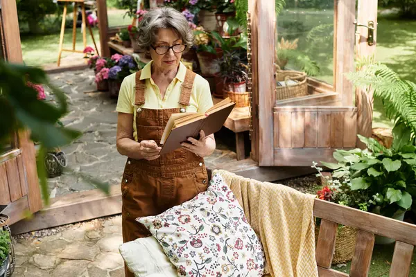 A beautiful senior woman stands in her lush garden, immersed in a book amidst colorful flowers. — Stock Photo