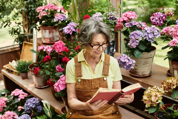 Senior woman in glasses relaxes in a garden, reading a book among colorful flowers. — Stock Photo