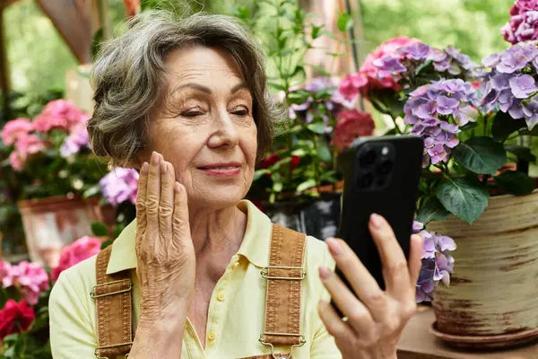 A joyful elderly woman admires her smartphone while surrounded by colorful garden flowers. — Stock Photo