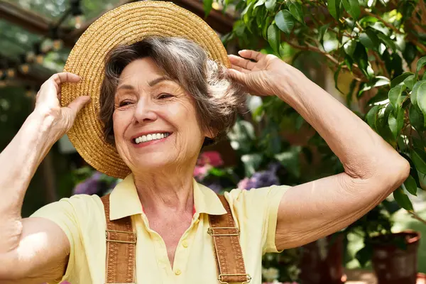A senior woman enjoys the beauty of her colorful garden, smiling as she adjusts her sunhat. — Stock Photo