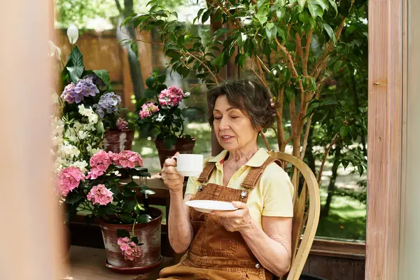 A serene senior woman enjoys tea in her garden, surrounded by vibrant flowers. — Stock Photo