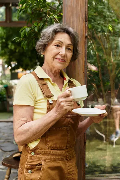 A beautiful senior woman stands in her lush garden, savoring a moment with a warm cup of tea. — Stock Photo