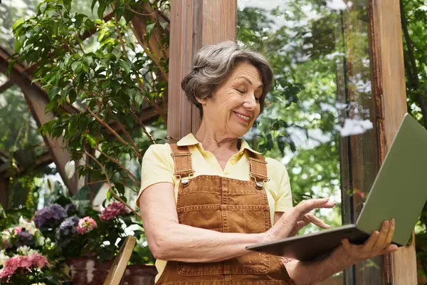 A senior woman smiles at her laptop, surrounded by blooming flowers in a sunny garden. — Stock Photo
