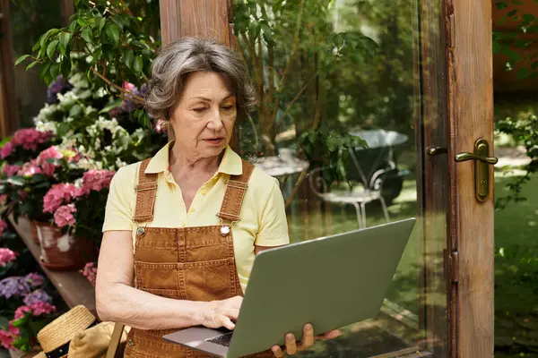 A beautiful senior woman engages with her laptop amidst vibrant flowers in a serene garden setting. — Stock Photo