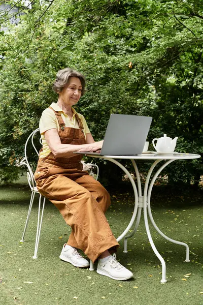 A beautiful senior woman sits at a garden table, focused on her laptop under lush greenery. — Stock Photo