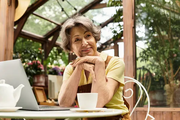 A beautiful senior woman relaxes in a lush garden, sipping tea while engaging with her laptop. — Stock Photo