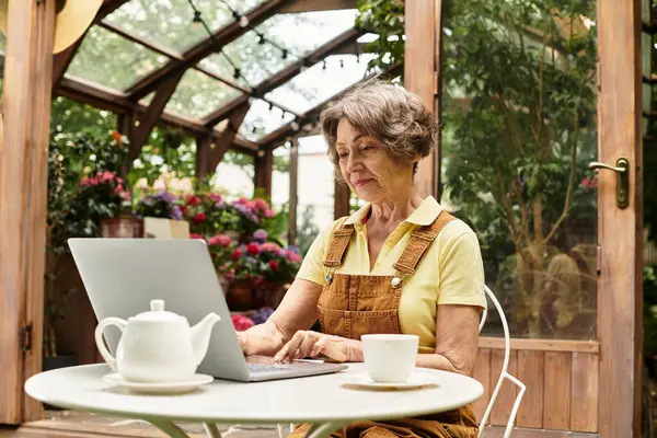 A beautiful senior woman sits at a table in a garden, focused on her laptop while sipping tea. — Stock Photo