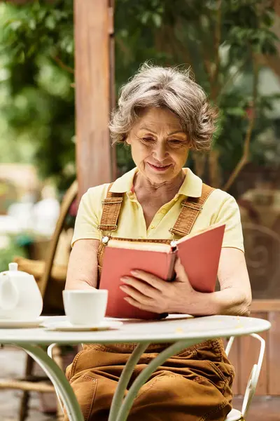 A beautiful senior woman sits at a table, absorbed in her book, surrounded by greenery and flowers. — Stock Photo