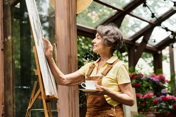 Senior woman engaged in painting in a lovely garden while savoring a cup of tea in peace. — Stock Photo