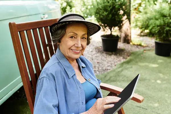 Smiling woman relaxes in a garden chair, using a tablet with vibrant greenery around her. — Stock Photo