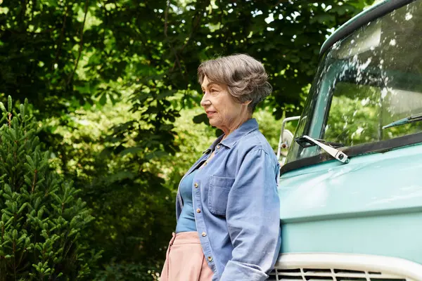 A senior woman smiles beside a vintage car as sunlight filters through the leaves. — Stock Photo
