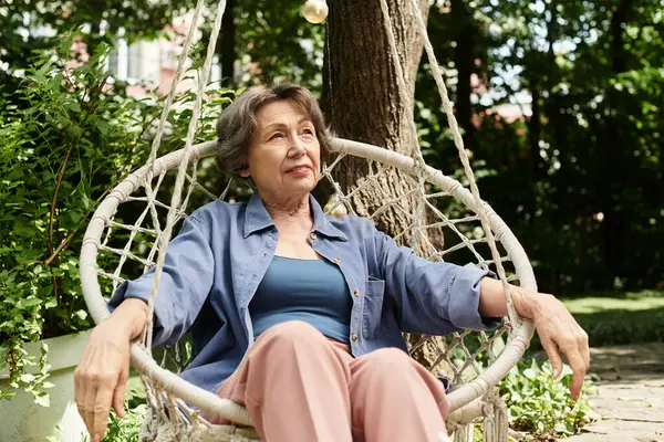 A serene senior woman relaxes on a garden swing amid lush greenery and sunlight. — стоковое фото
