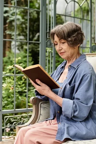 A beautiful senior woman relaxes in a serene garden while deeply engaged in a book on a sunny day. — Stock Photo
