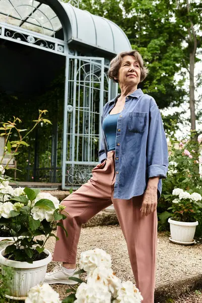 A senior woman stands gracefully among blooming flowers, soaking up the sun in her lush garden. — Stock Photo