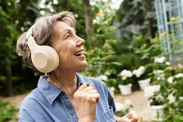 Senior woman with headphones dances happily among blooming flowers in a lush garden. — стокове фото