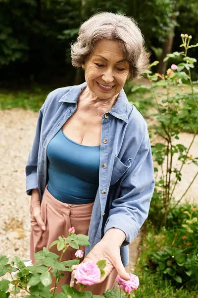Senior woman smiles joyfully while tending to pink roses in a lush garden during daylight. — Stock Photo