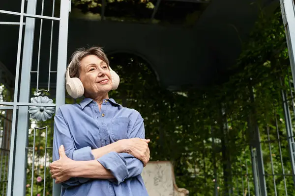 A senior woman stands peacefully in a lush garden wearing headphones, embracing natures beauty. — Stock Photo
