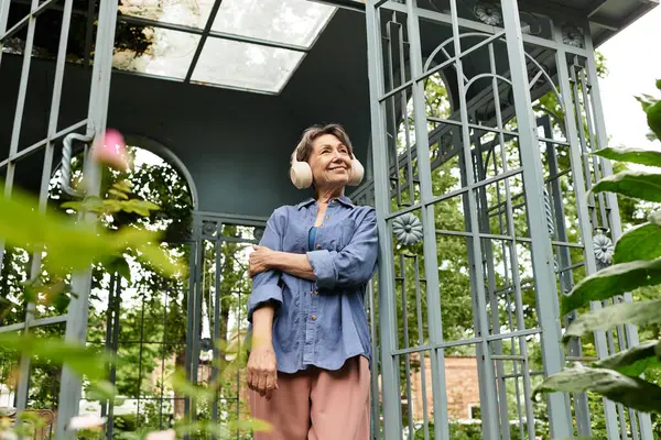 A cheerful senior woman in a cozy garden gazebo embraces tranquility surrounded by lush greenery. — Stock Photo