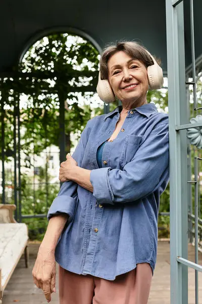 Beautiful senior woman relaxes in a serene garden, wearing headphones and smiling warmly. — Stock Photo