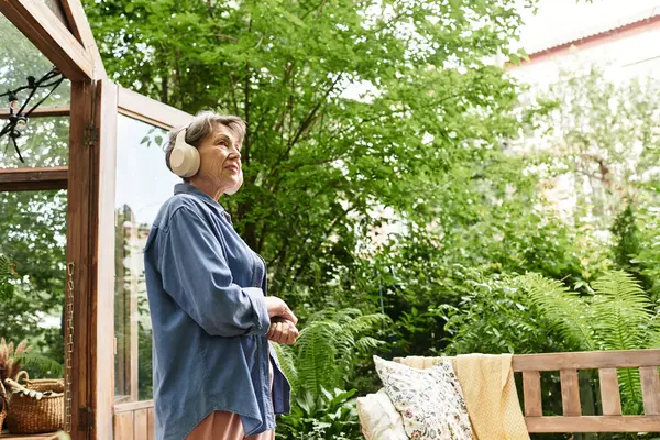 A senior woman enjoys music in her lush garden, surrounded by greenery and cozy seating. — Stock Photo