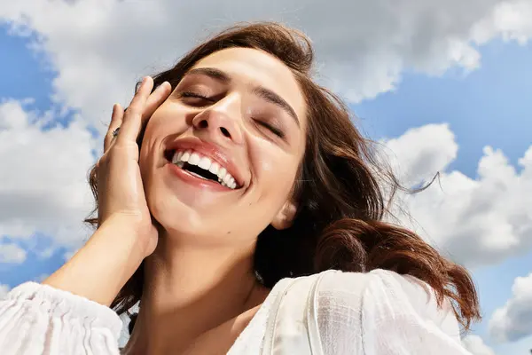 A young woman with wavy brown hair smiles brightly while enjoying a sunny day outdoors with clouds. — Stock Photo