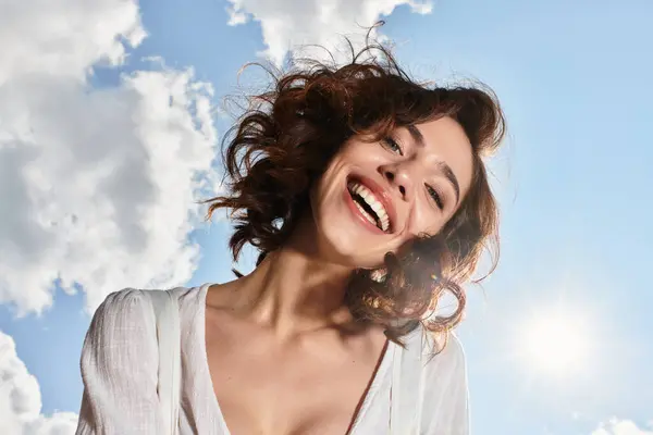 A young woman with wavy brown hair smiles joyfully under a clear blue sky on a summer day. — Stock Photo