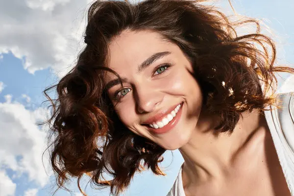 A pretty young woman with wavy brown hair smiles joyfully under a vibrant summer sky outdoors. — Stock Photo