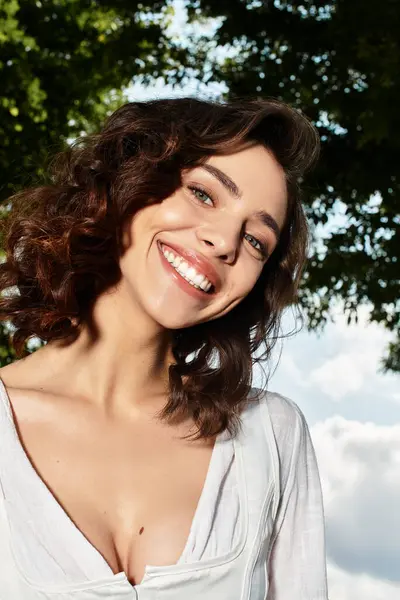 A young woman with wavy brown hair smiles brightly while standing outdoors on a summer day. — Stock Photo