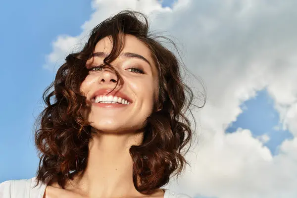A joyful young woman with wavy brown hair smiles brightly, embracing the summer day outdoors. — Stock Photo