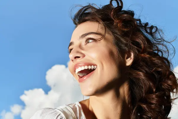A young woman with wavy brown hair smiles brightly against a clear blue sky on a sunny day. — Stock Photo