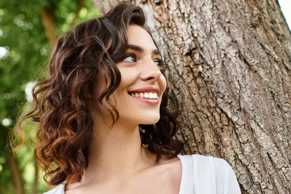 A pretty woman with wavy brown hair smiles while standing next to a tree in summer warmth. — Stock Photo