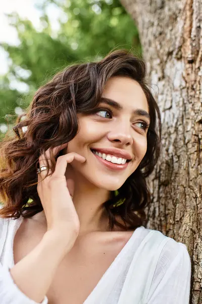 A pretty young woman with wavy brown hair smiles happily while enjoying a summer day outdoors. — Stock Photo