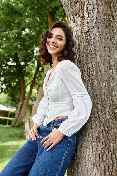 A young woman with wavy brown hair smiles joyfully while leaning against a tree on a summer day. — Stock Photo
