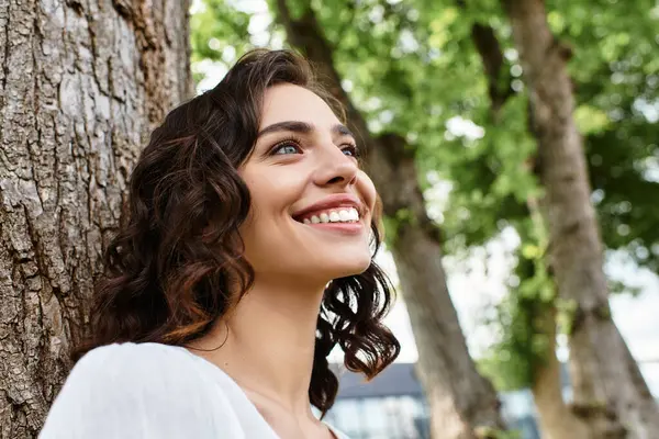 A young woman with wavy brown hair smiles joyfully while enjoying a sunny day in the park. — Stock Photo
