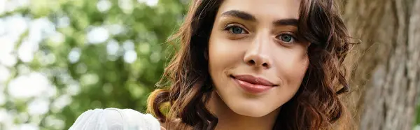 A pretty young woman with wavy brown hair smiles warmly in the summer sunshine among green trees, banner — Stock Photo