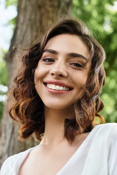 Wavy brown-haired woman smiles brightly under the sun in a peaceful outdoor park setting. — Stock Photo