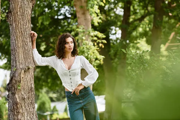 A pretty young woman with wavy brown hair leans against a tree in a lush park on a sunny day. — Stock Photo