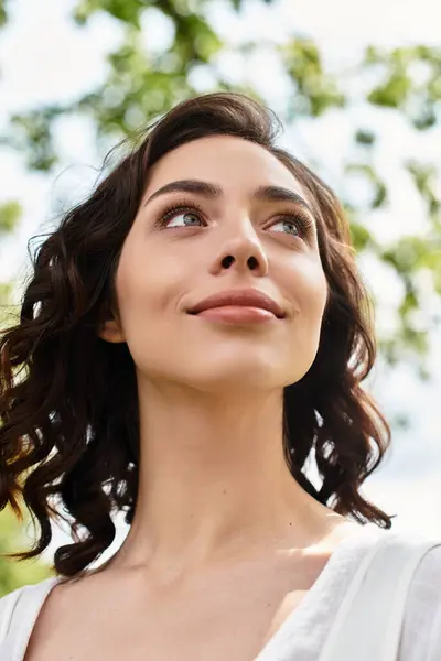 Pretty young woman with wavy brown hair looks up, enjoying the warm sunshine in a summer park. — Stock Photo