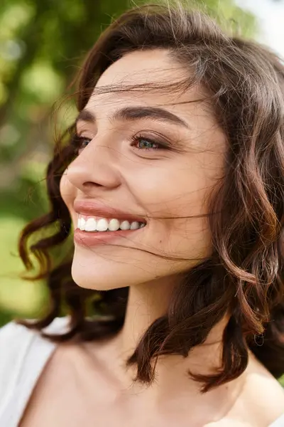 A young woman with wavy brown hair smiles warmly while enjoying a sunny day in the park. — Stock Photo
