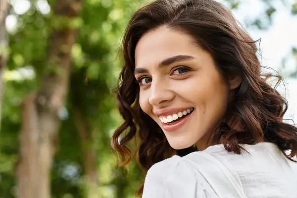A cheerful young woman with wavy brown hair enjoys a sunny summer day in a lush park. — Stock Photo