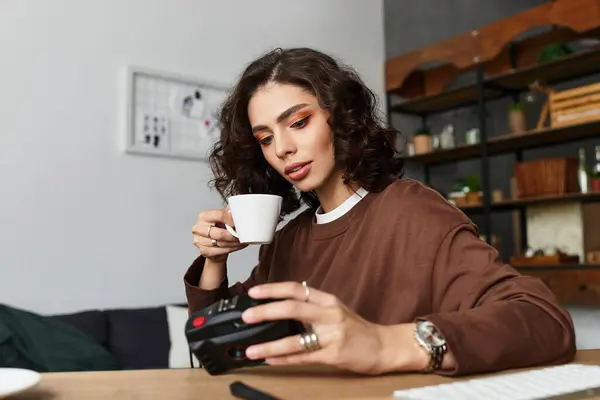 Young woman with curly hair sips coffee and checks her camera in a cozy home office setup. — Stock Photo
