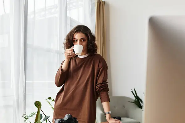 A beautiful young woman sips coffee while capturing moments in her cozy home workspace. — Stock Photo
