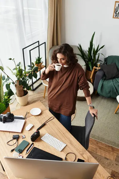 Curly-haired woman sips coffee at her desk while pursuing freelance photography during the day. — Stock Photo