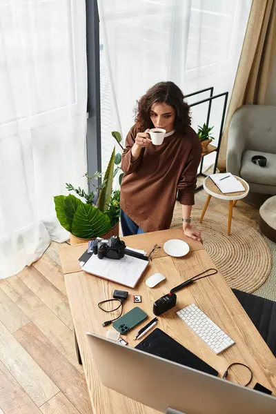A young woman with curly hair drinks coffee while working in her plant-filled home office. — Stock Photo